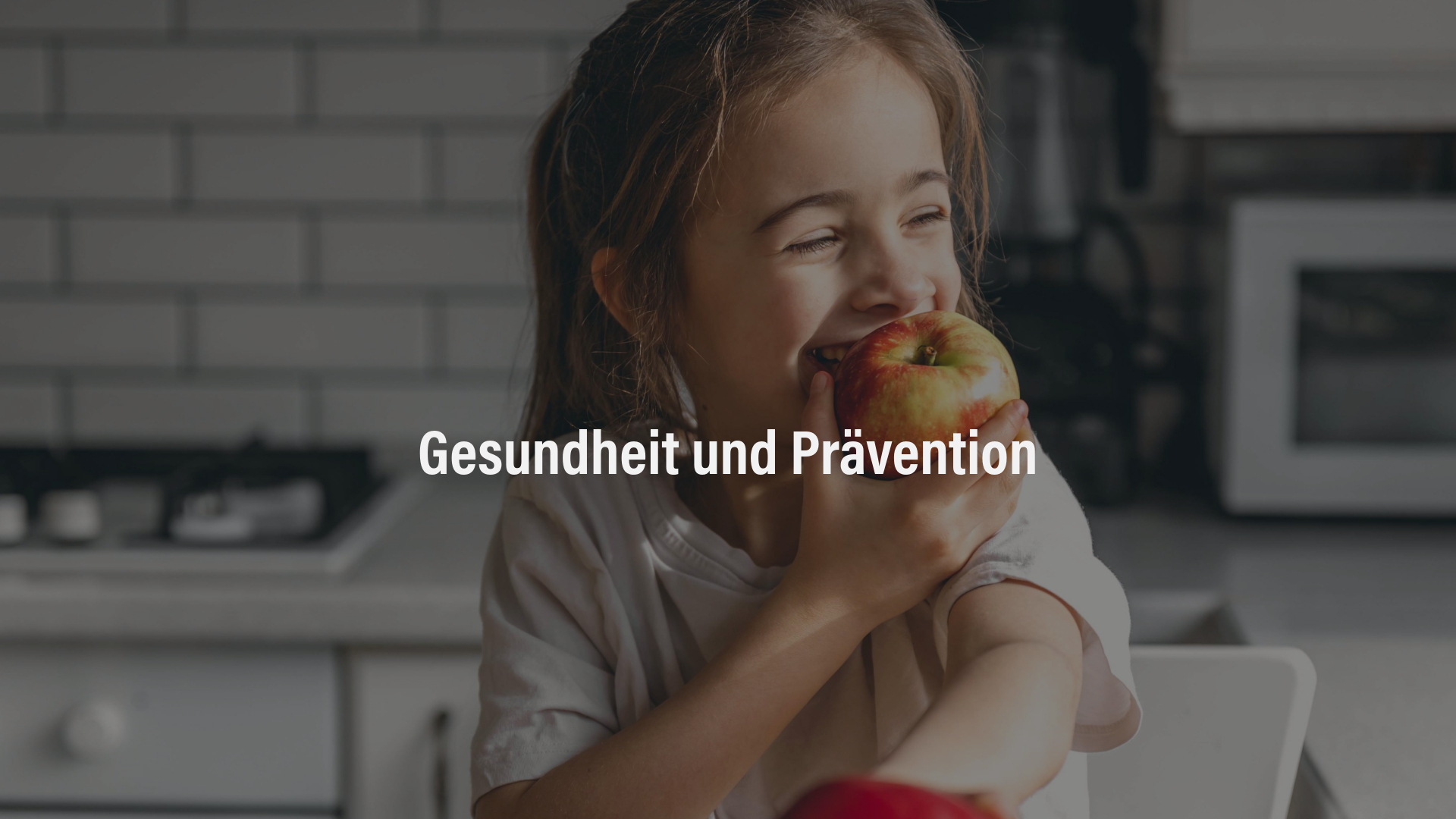 Ein lachendes Mädchen sitzt in der Küche und beißt in einen Apfel, während frisches Obst auf dem Tisch liegt.