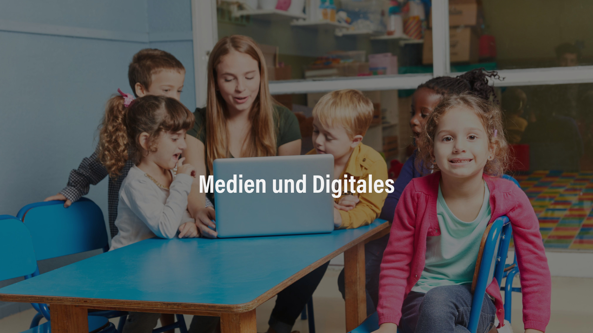 Medien und Digitales Eine junge Frau sitzt mit Kindern an einem blauen Tisch und zeigt ihnen etwas auf einem Laptop.