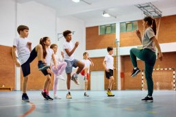 Mehrere Kinder und eine Trainerin üben gemeinsam eine Bewegungseinheit in einer Sporthalle.