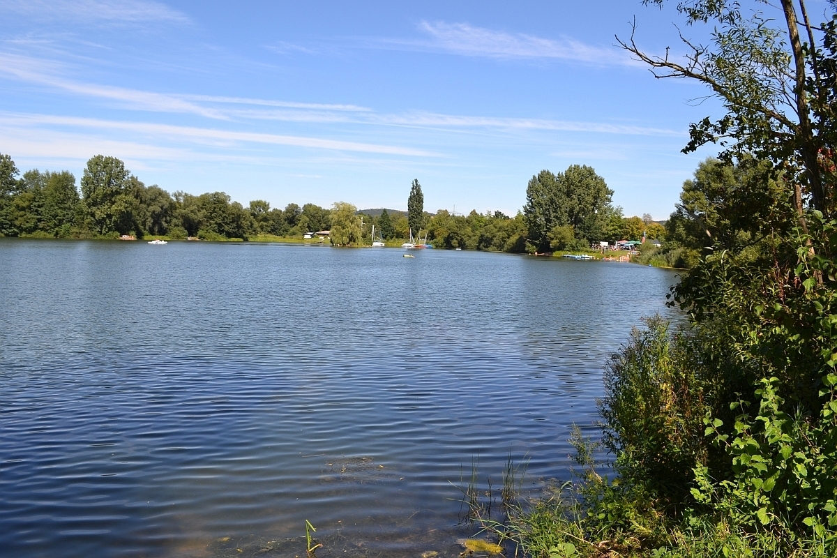 Blick auf einen See. Im Hintergrund das andere Ufer mit vielen Bäumen und ein blauer Himmel mit Schleierwolken. Auf dem See sind weit hinten kleine weiße Boote zu erkennen. Rechts sind grüne Büsche und Hecken, weiter hinten rechts lässt sich ein Campingplatz erkennen.
