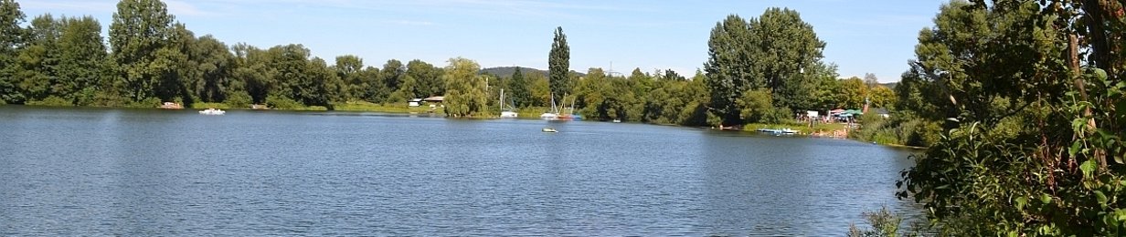 Blick auf einen See. Im Hintergrund das andere Ufer mit vielen Bäumen und ein blauer Himmel mit Schleierwolken. Auf dem See sind weit hinten kleine weiße Boote zu erkennen. Rechts sind grüne Büsche und Hecken, weiter hinten rechts lässt sich ein Campingplatz erkennen.