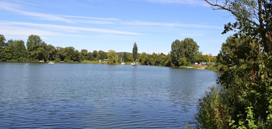 Blick auf einen See. Im Hintergrund das andere Ufer mit vielen Bäumen und ein blauer Himmel mit Schleierwolken. Auf dem See sind weit hinten kleine weiße Boote zu erkennen. Rechts sind grüne Büsche und Hecken, weiter hinten rechts lässt sich ein Campingplatz erkennen.