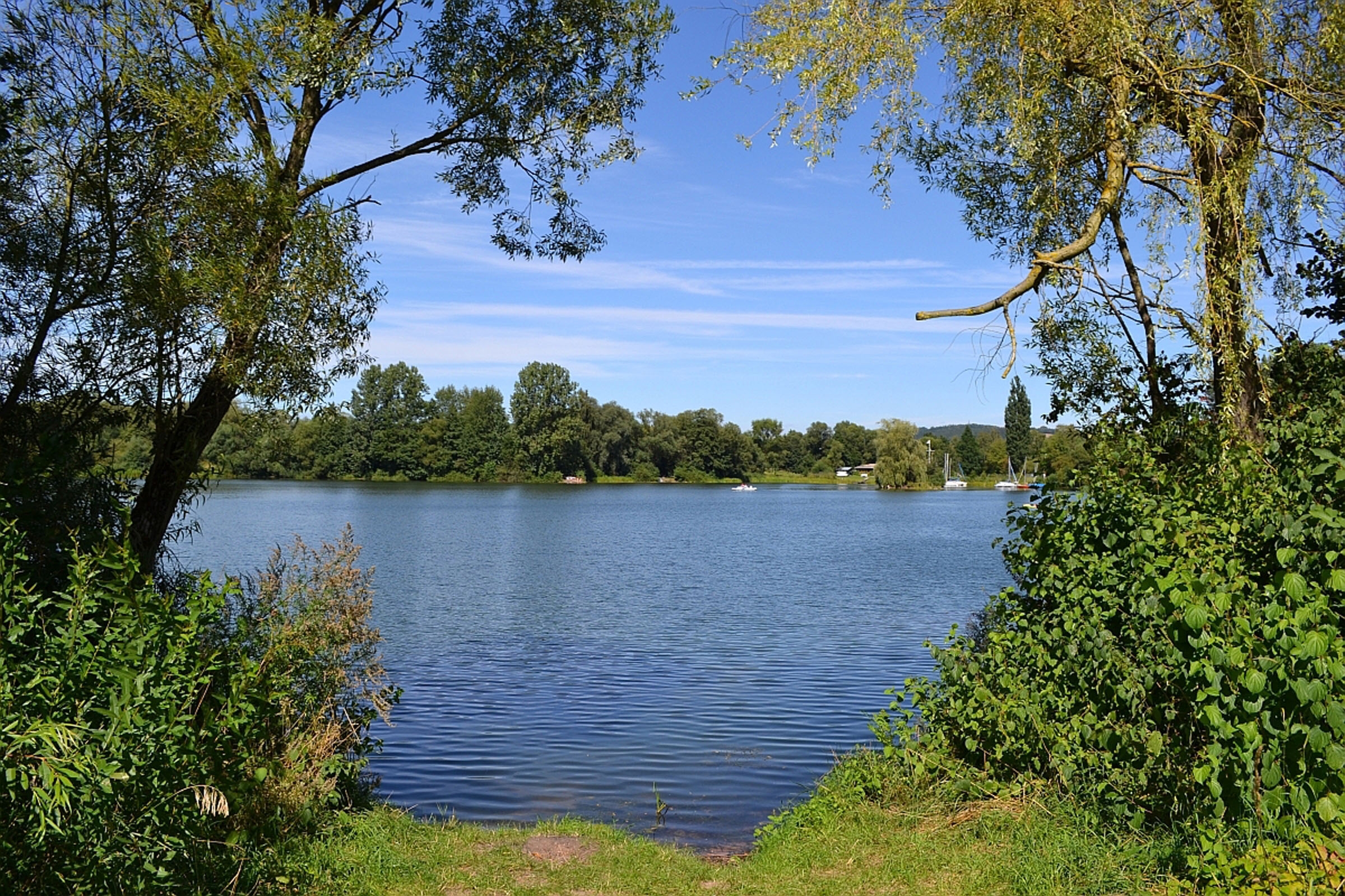 Blick zwischen zwei Bäumen und Hecken auf einen See. Im Hintergrund das andere Ufer mit vielen Bäumen und ein blauer Himmel mit Schleierwolken. Auf dem See sind weit hinten kleine weiße Boote zu erkennen.