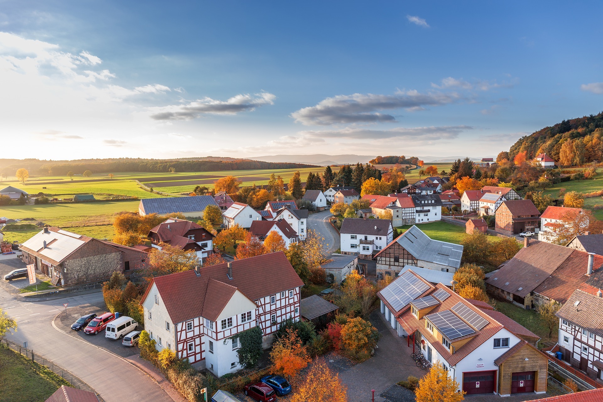 Luftbild von einem Dorf in einer Herbstlandschaft. Zu sehen sind Bäume mit bunten Blättern zwischen den Häusern, Im Hintergrund grüne Wiesen und Wald, blauer Himmel mit ein paar Wolken.
