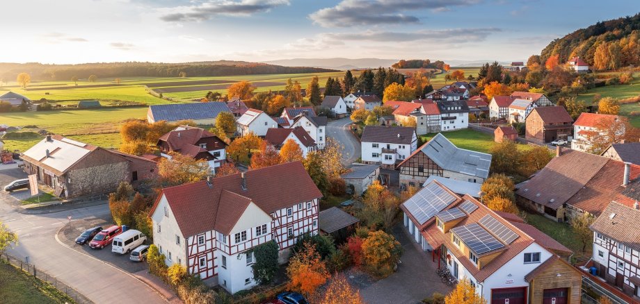Luftbild von einem Dorf in einer Herbstlandschaft. Zu sehen sind Bäume mit bunten Blättern zwischen den Häusern, Im Hintergrund grüne Wiesen und Wald, blauer Himmel mit ein paar Wolken.