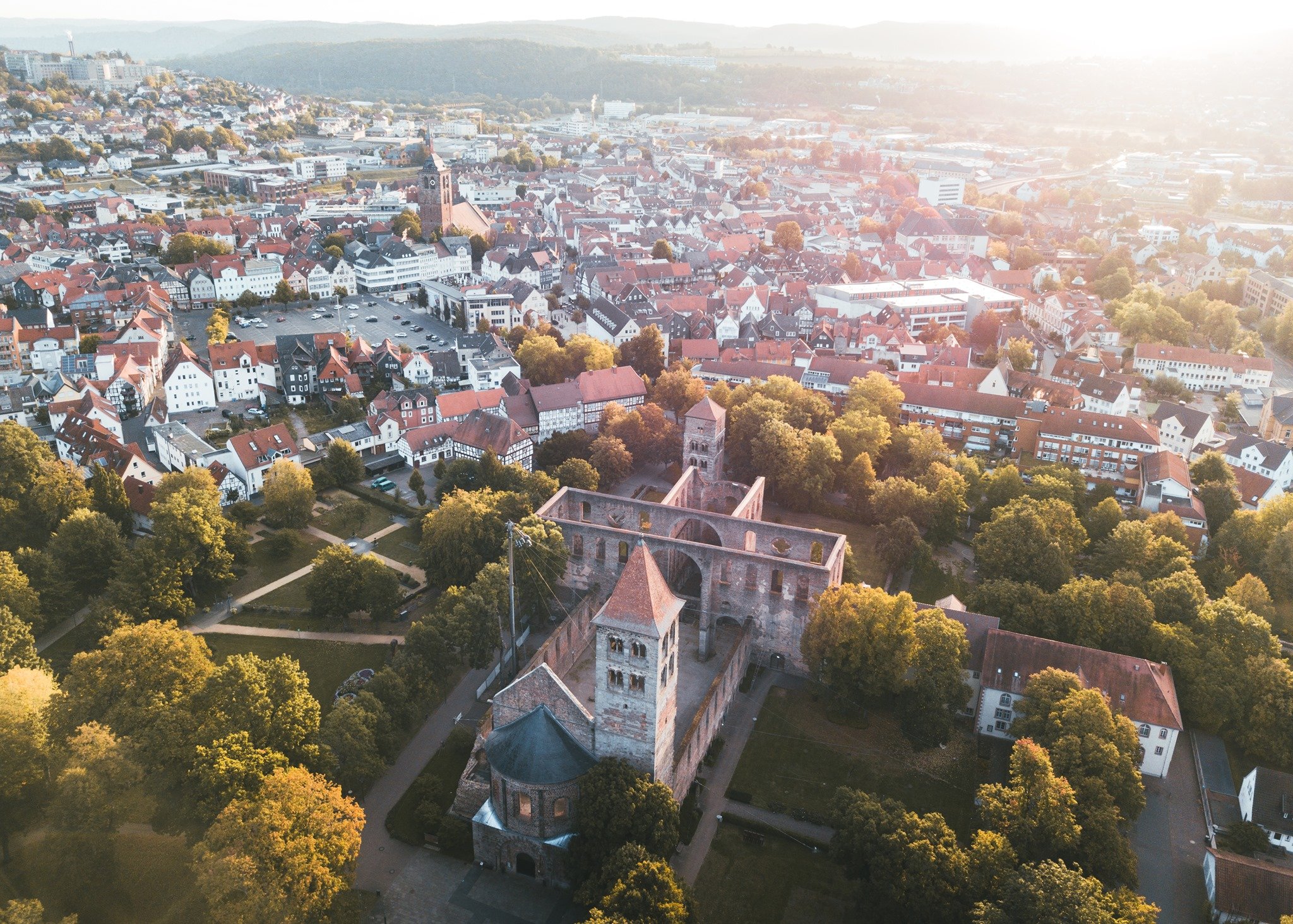 In Abendsonne ein Luftbild über die Stadt Bad Hersfeld, im Fokus ist die Stiftsruine mit dem Park Stiftsbezirk mit braunen Gehwegen, dunkelgrünem Gras und herbstlich gefärbten Bäumen. Im Hintergrund die Stadt mit Stadtkirche, Marktplatz, Häusern, Klinikum und weiter hinten Bergen mit Wald.