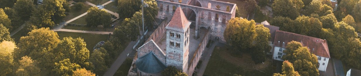 In Abendsonne ein Luftbild über die Stadt Bad Hersfeld, im Fokus ist die Stiftsruine mit dem Park Stiftsbezirk mit braunen Gehwegen, dunkelgrünem Gras und herbstlich gefärbten Bäumen. Im Hintergrund die Stadt mit Stadtkirche, Marktplatz, Häusern, Klinikum und weiter hinten Bergen mit Wald.