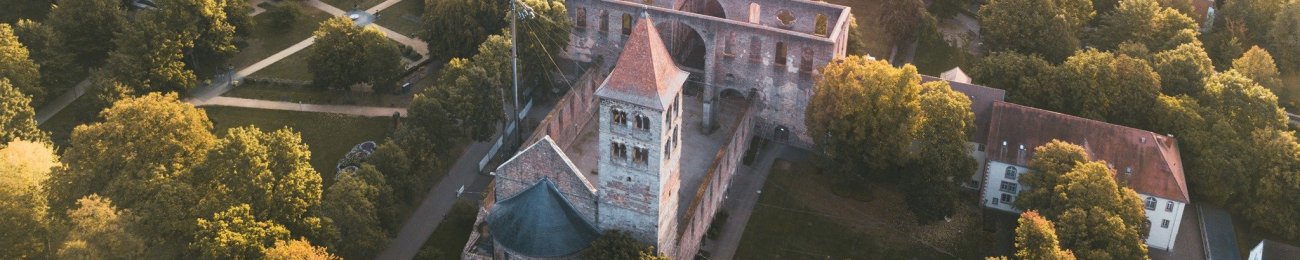 In Abendsonne ein Luftbild über die Stadt Bad Hersfeld, im Fokus ist die Stiftsruine mit dem Park Stiftsbezirk mit braunen Gehwegen, dunkelgrünem Gras und herbstlich gefärbten Bäumen. Im Hintergrund die Stadt mit Stadtkirche, Marktplatz, Häusern, Klinikum und weiter hinten Bergen mit Wald.