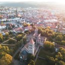 In Abendsonne ein Luftbild über die Stadt Bad Hersfeld, im Fokus ist die Stiftsruine mit dem Park Stiftsbezirk mit braunen Gehwegen, dunkelgrünem Gras und herbstlich gefärbten Bäumen. Im Hintergrund die Stadt mit Stadtkirche, Marktplatz, Häusern, Klinikum und weiter hinten Bergen mit Wald.
