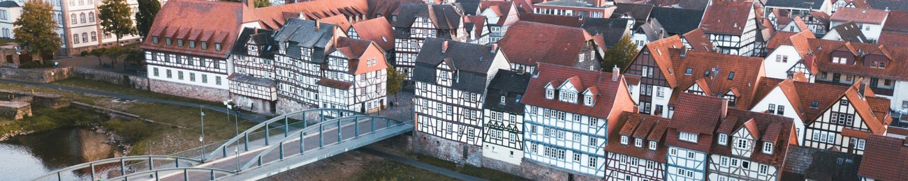 Luftbild über die Stadt Rotenburg an der Fulda mit Blick auf die Fulda, die alte Fuldabrücke und die Fachwerkhäuser der Innenstadt. Im Hintergrund ist ein bewaldeter Berg zu sehen. 
