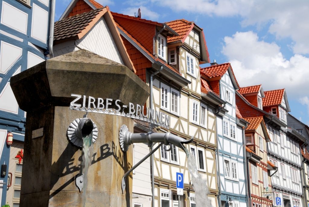 Zu sehen ist ein Betonpfeiler als Brunnen. Von ihm gehen zwei silberne Rohre, aus denen Wasser fließt. Auf den Rohren ist das Wort "Zirbes-Brunnen" aus Metall angebracht. Im Hintergrund sind Fassaden bunter Fachwerkhäuser zu sehen sowie ein blauer Himmel mit weißen Wolken. 