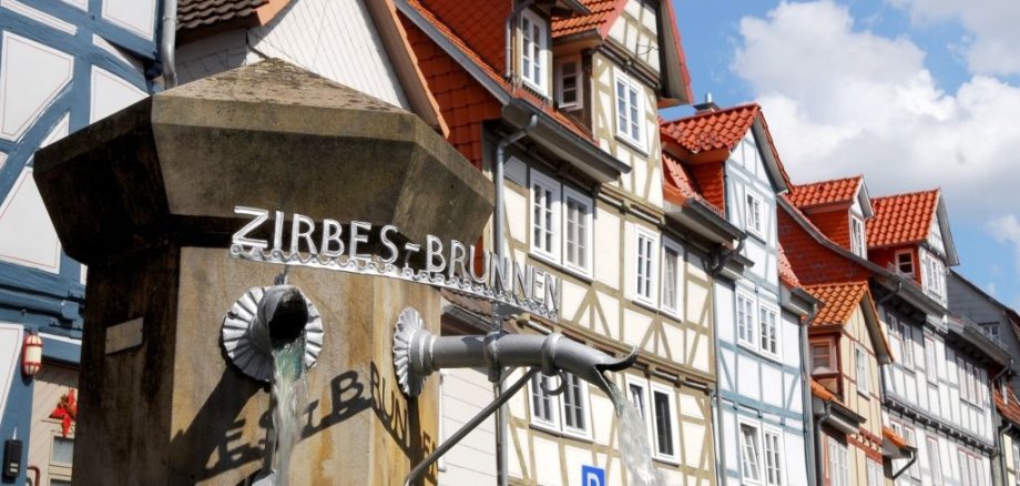 Zu sehen ist ein Betonpfeiler als Brunnen. Von ihm gehen zwei silberne Rohre, aus denen Wasser fließt. Auf den Rohren ist das Wort "Zirbes-Brunnen" aus Metall angebracht. Im Hintergrund sind Fassaden bunter Fachwerkhäuser zu sehen sowie ein blauer Himmel mit weißen Wolken. 