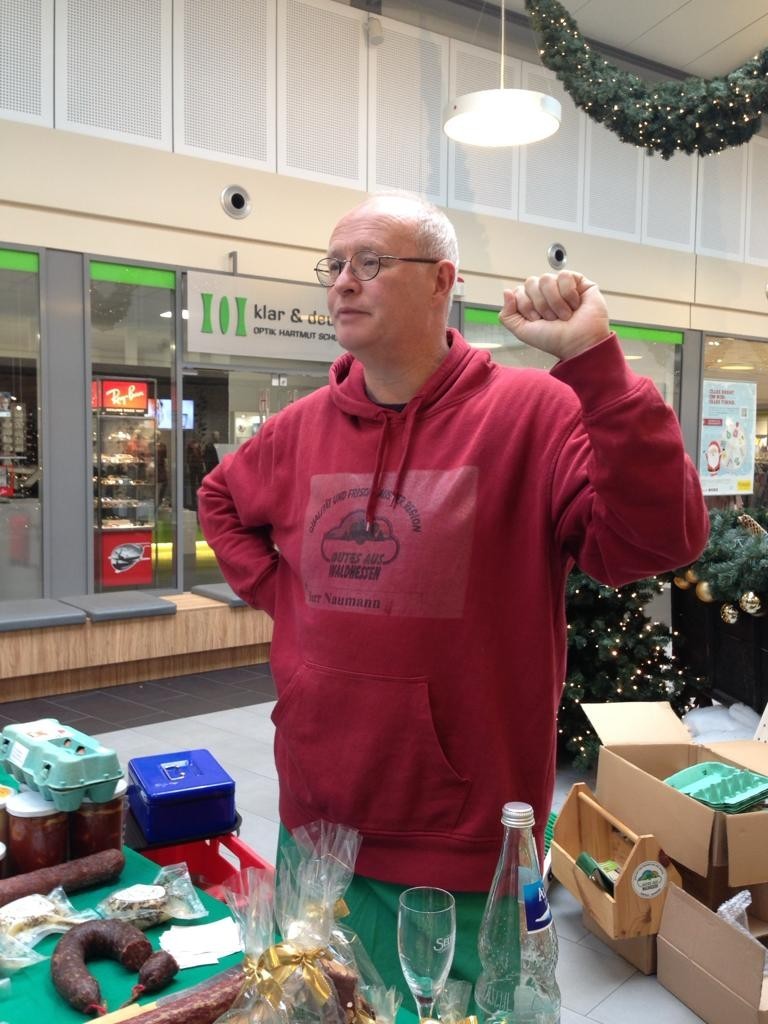 Ein Mann in einem roten Pullover steht in einem Einkaufszentrum. Auf dem Tisch vor ihm liegen Stracken, Eier, eine Kasse und ein Glas. Im Hintergrund sieht man Weihnachtsdekoration.