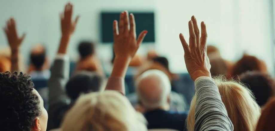 Eine Gruppe von Menschen in einem Sitzungssaal hebt die Hände zur Abstimmung. Die Teilnehmer sind von hinten zu sehen, der Raum ist gut beleuchtet, und der Fokus liegt auf den gehobenen Händen.