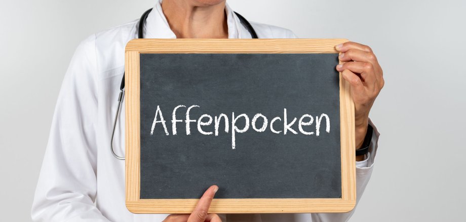 Arzt hält ein Schild hoch. Dort ist mit Kreide das Wort Affenpocken draufgeschrieben.