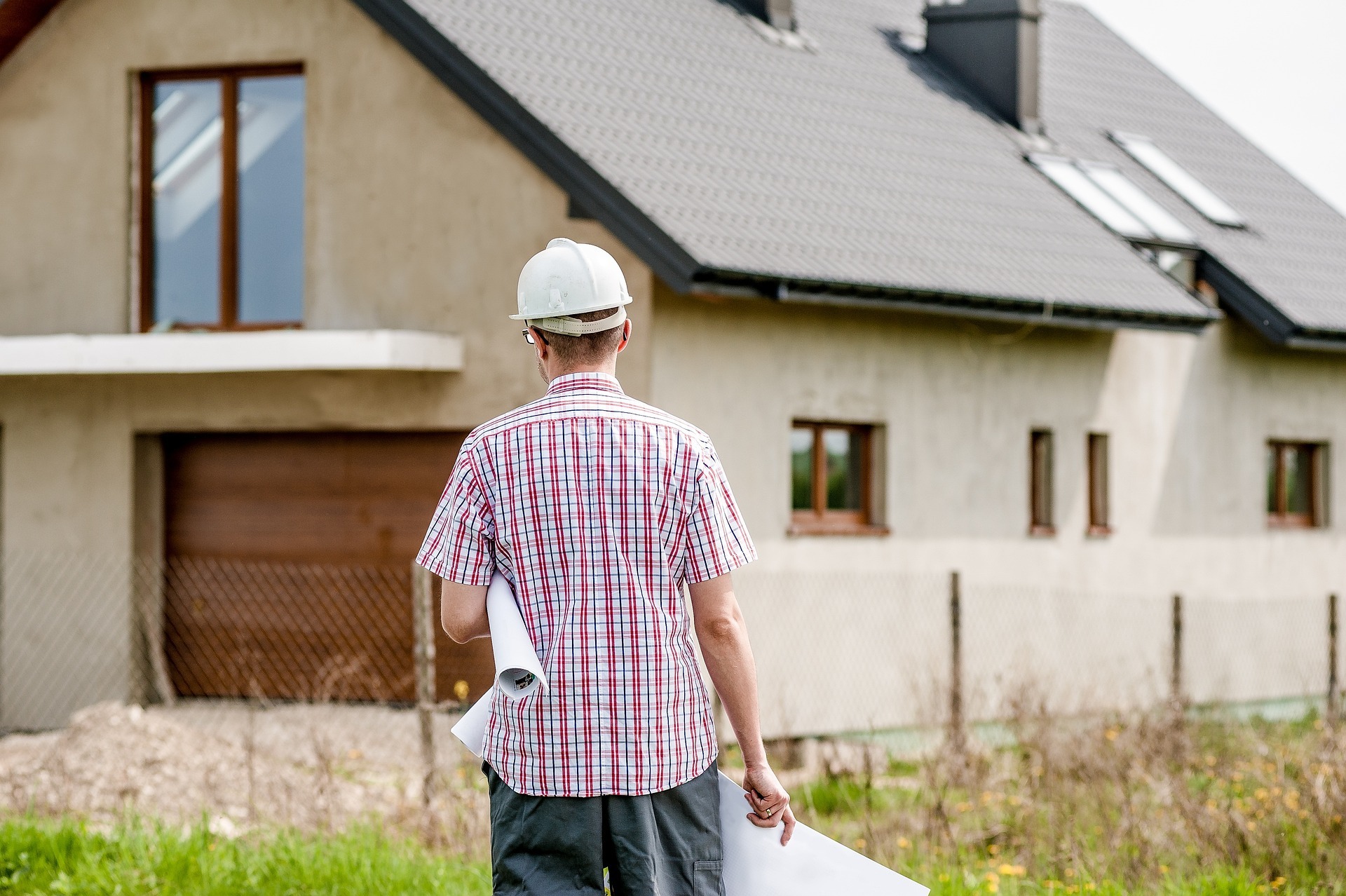 Rückansicht eines Mannes in Karohemd mit weißem Bauhelm und Bauplänen in der Hand und unter dem Arm. Im Hintergrund ein Neubau eines Einfamilienhauses.