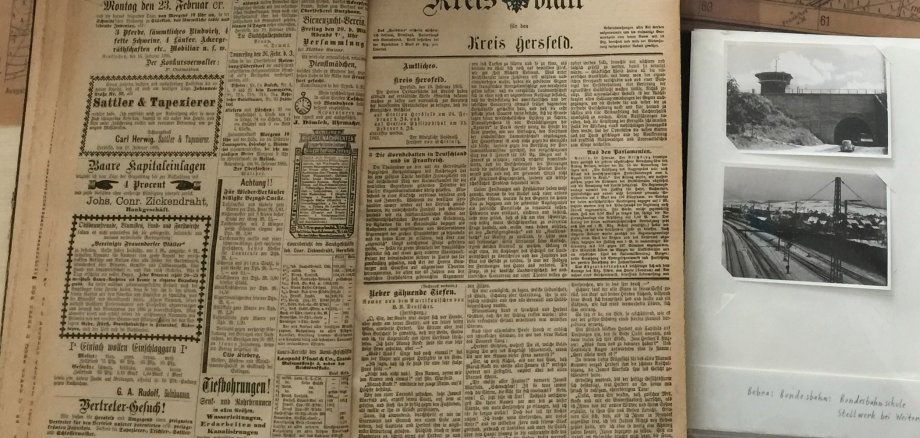 Links oben ein Karton, der halb geöffnet ist und worin alte Zeitungen liegen. Im Hintergrund liegt eine antike Karte, unten links eine historische Ausgabe des Kreisblatts von 1885. Rechts unten ein Fotoalbum mit schwarz-weiß Fotos vom Bahnhof Bebra und einem Tunnel. Oben in der Mitte liegt ein Dia, daneben steht Dorflinden.