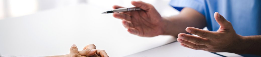 Nahaufnahme eines weißen Schreibtisches. Man sieht links verschränkte Hände, liegend auf dem Schreibtisch. Davor ein Formular auf einem Klemmbrett, daneben ein Stethoskop. Eine Person im blauen Kittel sitzt davor, die Hände nach vorne zeigend.