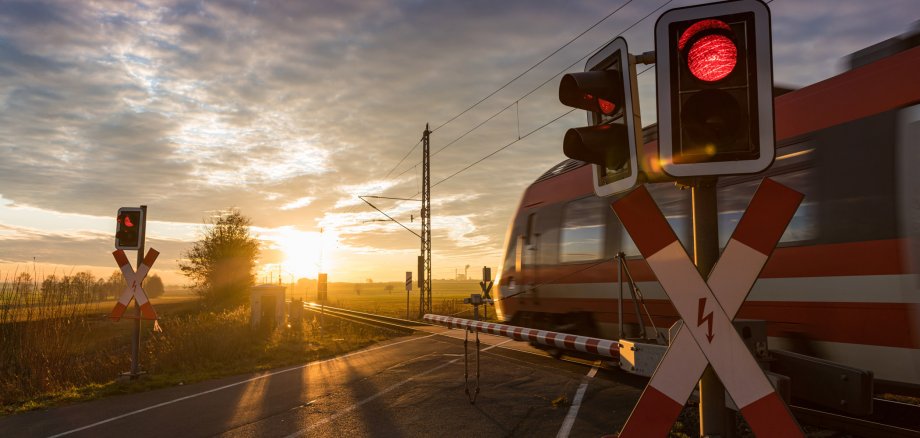 Bahnübergang im Sonnenuntergang. Schranken sind unten, ein roter Zug fährt durch.