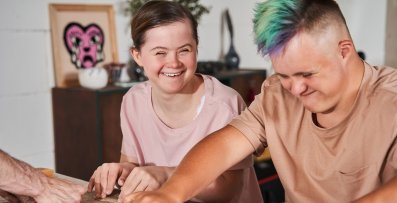 Portraitbild eines Mädchen und Jungens mit geistiger Behinderung. Sie lacht während sie den Jungen anschaut, der mit einem Nudelholz Ton auf einem Tisch ausrollt.