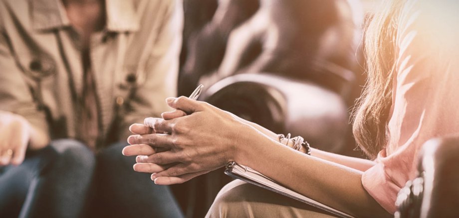 Nahaufnahme von zwei auf braunen Ledersesseln sitzenden Menschen. Zu sehen sind die Arme einer Frau, die ein Klemmbrett zwischen ihren Beinen und Armen lieen hat. Nach vorngebeugter Oberkörper. Die andere Person hat eine offene Hand.
