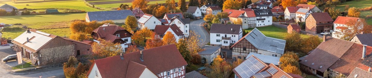 Luftbild von einem Dorf in einer Herbstlandschaft. Zu sehen sind Bäume mit bunten Blättern zwischen den Häusern, Im Hintergrund grüne Wiesen und Wald, blauer Himmel mit ein paar Wolken.