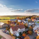Luftbild von einem Dorf in einer Herbstlandschaft. Zu sehen sind Bäume mit bunten Blättern zwischen den Häusern, Im Hintergrund grüne Wiesen und Wald, blauer Himmel mit ein paar Wolken.