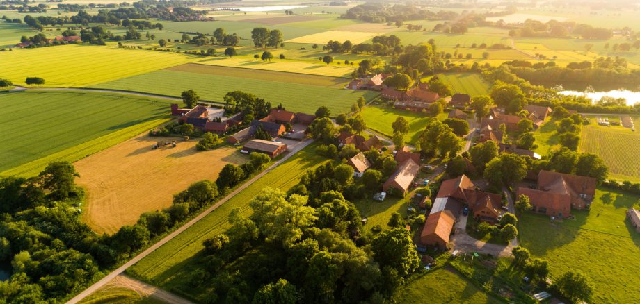 Einige Häuser eines Dorfes sind aus der Luft zu sehen. Darum sind viele Wiesen und Felder. Es sind viele Bäume zu sehen. Die Sonne geht gerade unter.
