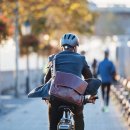 Eine Rückansicht eines Geschäftsmannes mit Fahrradhelm und lederner Umhängetasche auf einem Elektrofahrrad. Er ist auf einem Platz unterwegs, links verschwommen sind Bäume, rechts Stühle und weitere Menschen im Hitnergrund.