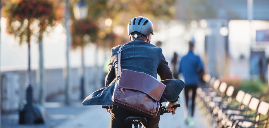 Eine Rückansicht eines Geschäftsmannes mit Fahrradhelm und lederner Umhängetasche auf einem Elektrofahrrad. Er ist auf einem Platz unterwegs, links verschwommen sind Bäume, rechts Stühle und weitere Menschen im Hitnergrund.