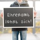 Ein junger Mann hält ein Schild hoch. Auf dem Schild steht Ehrenamt lohnt sich. 