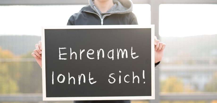 Ein junger Mann hält ein Schild hoch. Auf dem Schild steht Ehrenamt lohnt sich.