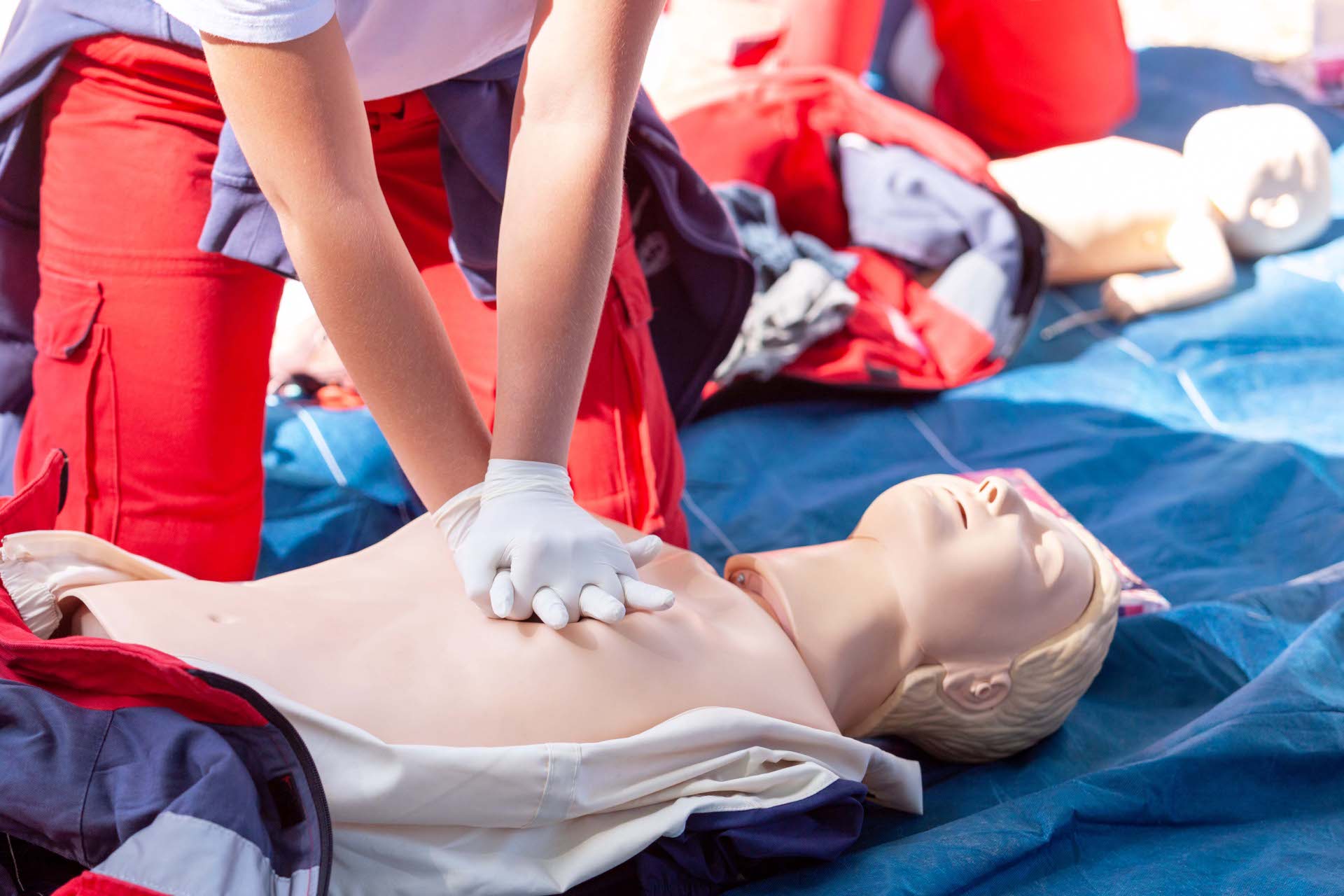 Eine Person in roter Einsatzkleidung und weißen Handschuhen führt Herzdruckmassage an einer Übungspuppe durch. Im Hintergrund ist eine weitere Reanimationspuppe zu sehen.
