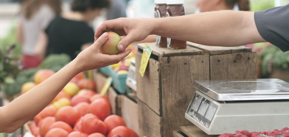 Die Hand eines Verkäufers überreicht der Hand eines Kunden einen Apfel. Im Hintergrund ist der Rest des Marktstandes mit vielen Äpfeln und einer Waage zu sehen.