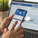 Zwei Hände, die ein Handy halten. Auf dem Handydisplay ist die Facebook-Registrierungsseite zu sehen. Im Hintergrund steht ein Laptop, hier ist ebenfalls die Facebook-Registrierungsseite zu sehen. Links neben dem Laptop steht zur Deko eine Blume und ein Mini-Einkaufswagen.