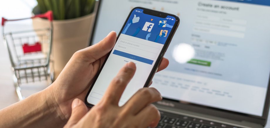 Zwei Hände, die ein Handy halten. Auf dem Handydisplay ist die Facebook-Registrierungsseite zu sehen. Im Hintergrund steht ein Laptop, hier ist ebenfalls die Facebook-Registrierungsseite zu sehen. Links neben dem Laptop steht zur Deko eine Blume und ein Mini-Einkaufswagen.
