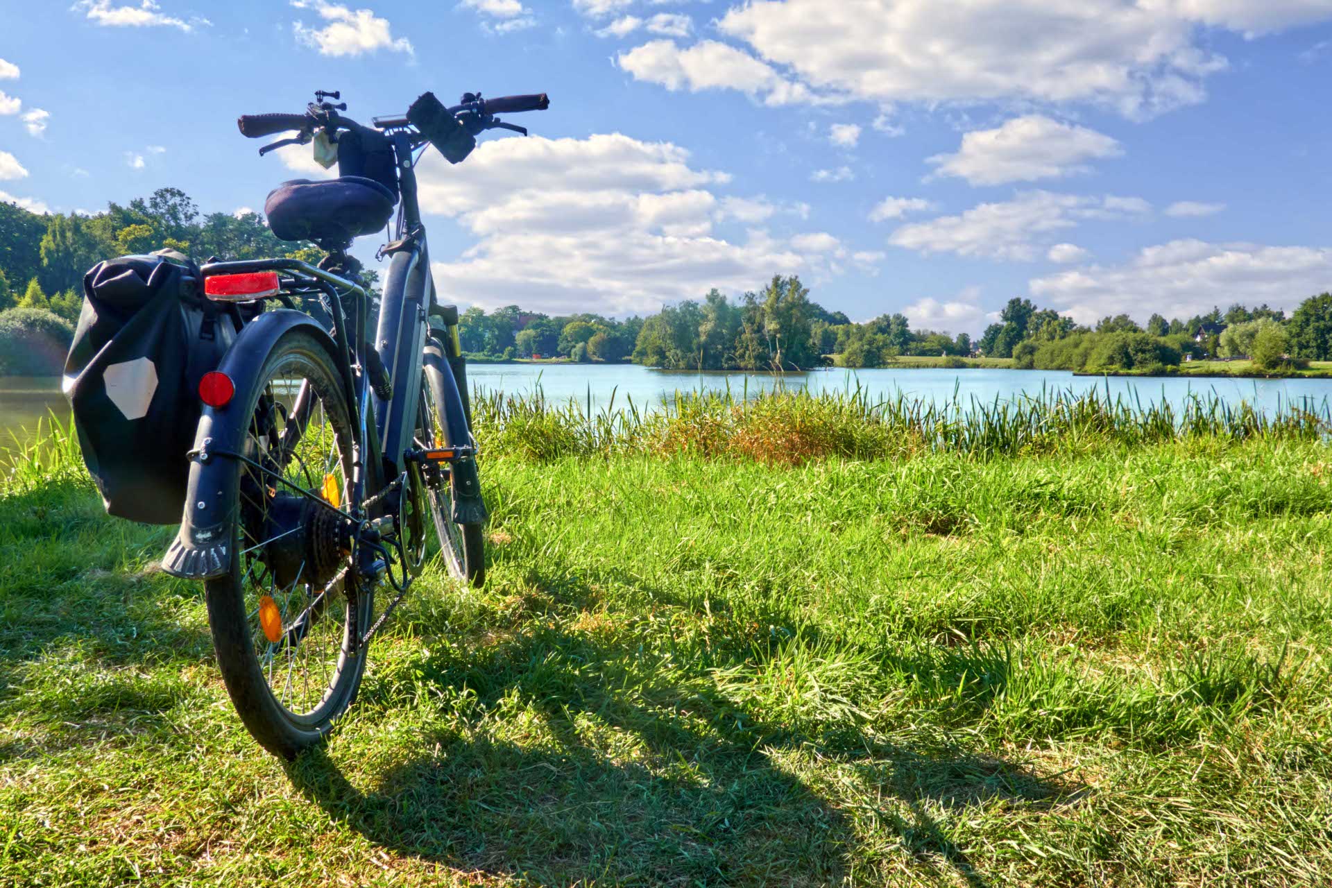 Ein schwarzes E-Bike mit Gepäcktasche steht auf einer grünen Wiese am Ufer eines Sees. Im Hintergrund ist eine bewaldete Uferlandschaft mit vereinzelten Häusern zu sehen. Der Himmel ist blau mit einigen weißen Wolken.