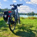 Ein schwarzes E-Bike mit Gepäcktasche steht auf einer grünen Wiese am Ufer eines Sees. Im Hintergrund ist eine bewaldete Uferlandschaft mit vereinzelten Häusern zu sehen. Der Himmel ist blau mit einigen weißen Wolken.