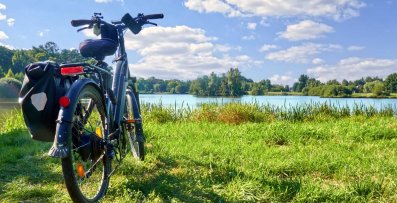 Ein schwarzes E-Bike mit Gepäcktasche steht auf einer grünen Wiese am Ufer eines Sees. Im Hintergrund ist eine bewaldete Uferlandschaft mit vereinzelten Häusern zu sehen. Der Himmel ist blau mit einigen weißen Wolken.