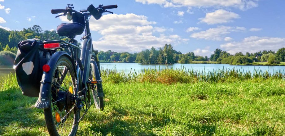 Ein schwarzes E-Bike mit Gepäcktasche steht auf einer grünen Wiese am Ufer eines Sees. Im Hintergrund ist eine bewaldete Uferlandschaft mit vereinzelten Häusern zu sehen. Der Himmel ist blau mit einigen weißen Wolken.