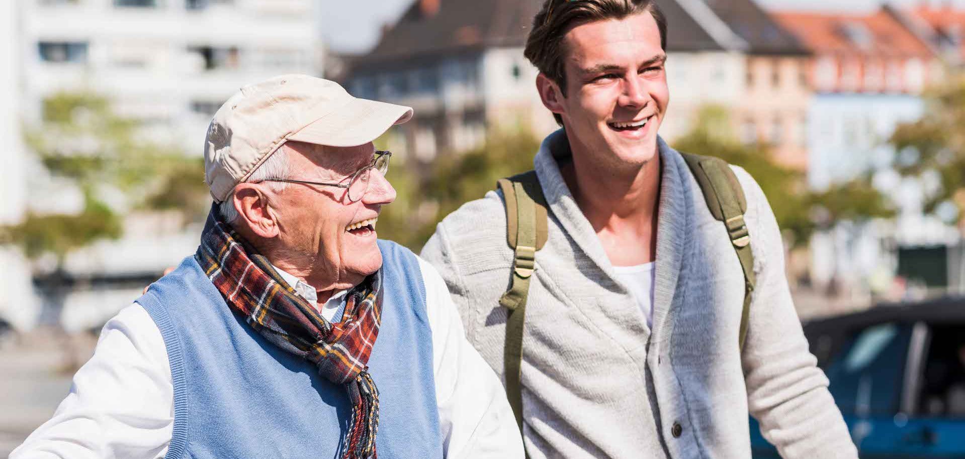 Ein älterer Mann mit Fahrrad und ein junger Mann mit Rucksack und Skateboard laufen lachend nebeneinander durch eine Ortschaft bei sonnigem Wetter.