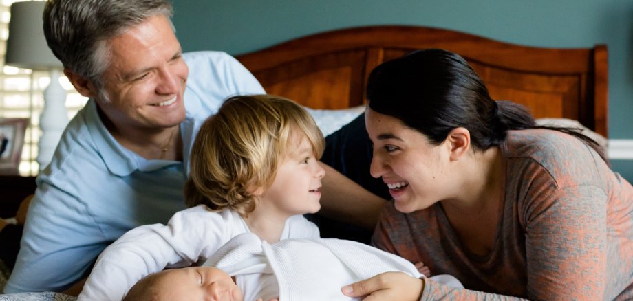 Lächelnde Eltern liegen auf einem Bett, zwischen ihnen liegt ein schlafendes Baby und ein kleiner Junge, der die Mutter anlächelt.  