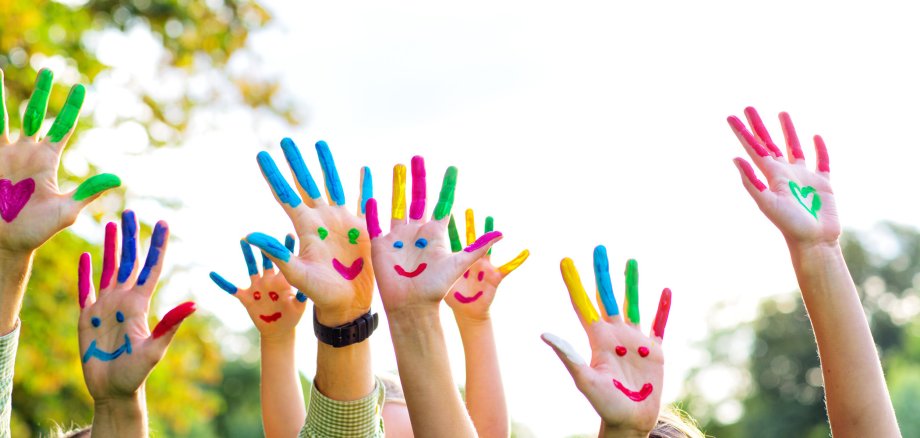 Viele hochgehaltene Arme mit Händen. Auf den Händen sind bunte Smileys gemalt, die Finger sind farbig gemalt. 
