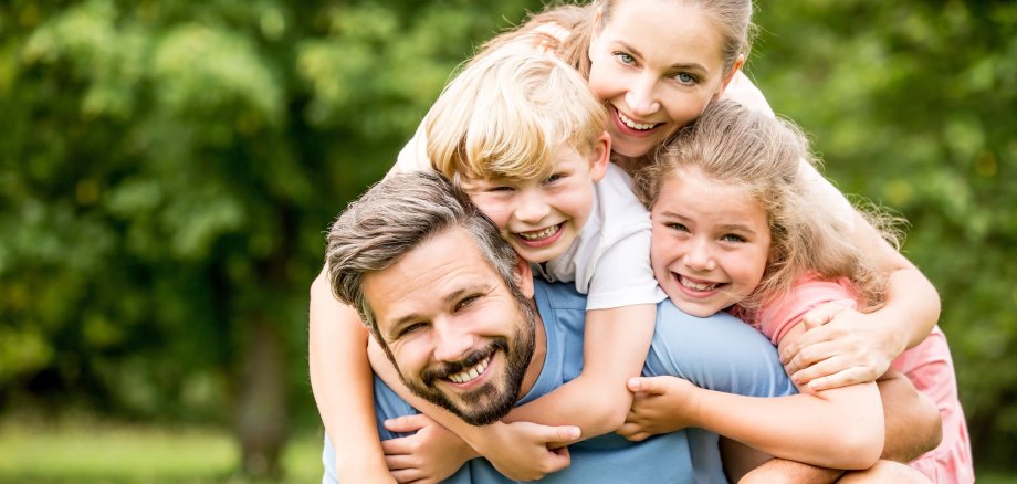 Nahaufnahme einer glücklichen Familie bestehend aus Mutter, Vater, Junge und Mädchen, die sich umarmend in die Kamera schauen. Weichgezeichneter Hintergrund mit Sommerwiese und Laubbäumen.