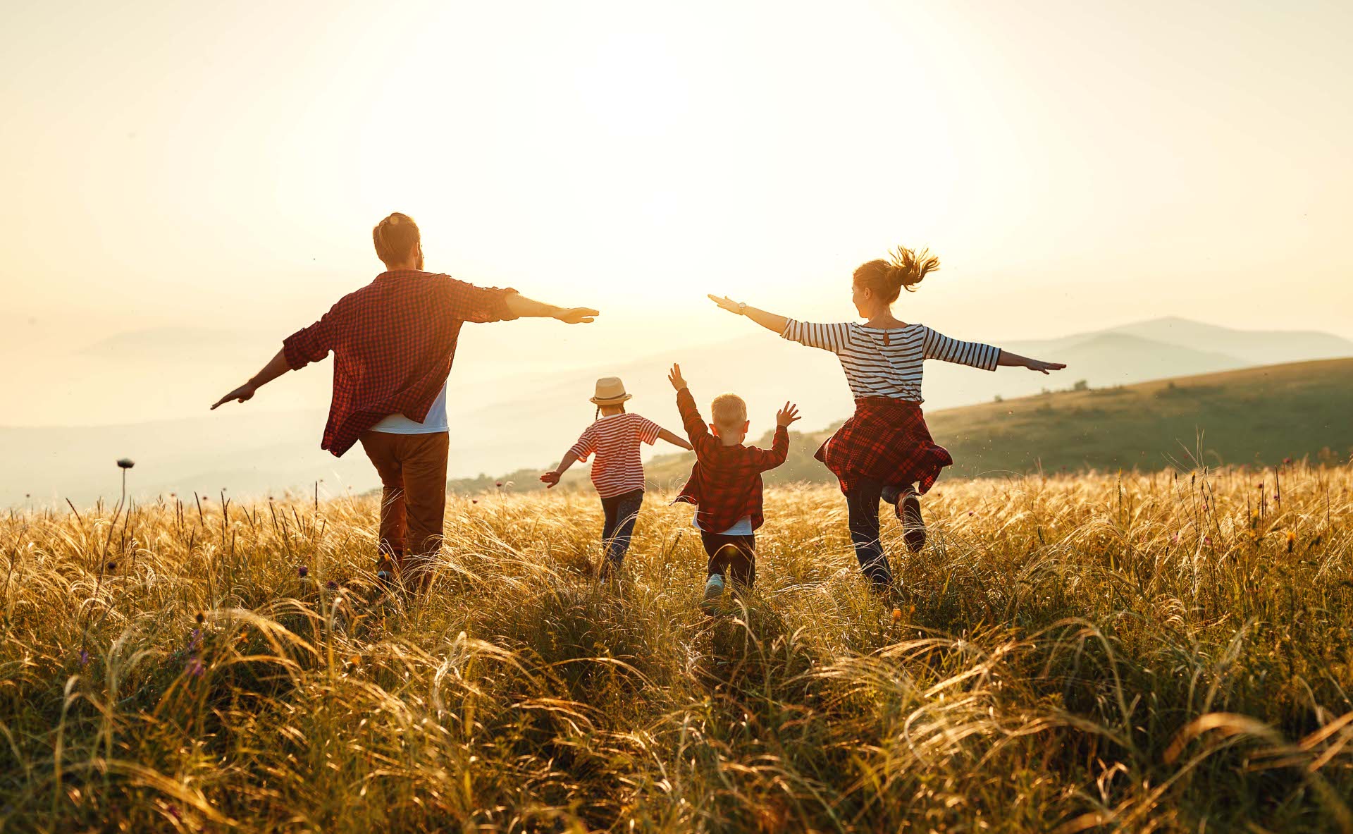 Eine Familie mit zwei Kindern läuft mit ausgebreiteten Armen durch ein Feld bei Sonnenuntergang. Alle sind von hinten zu sehen, im Hintergrund eine hügelige Landschaft.
