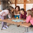 Eine Familie spielt in ihrem Wohnzimmer ein Spiel. Neben den beiden Eltern spielen auch drei Mädchen mit. Sie alle sind fröhlich.