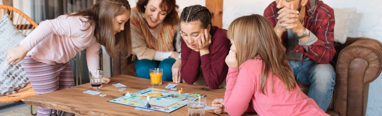 Eine Familie spielt in ihrem Wohnzimmer ein Spiel. Neben den beiden Eltern spielen auch drei Mädchen mit. Sie alle sind fröhlich.
