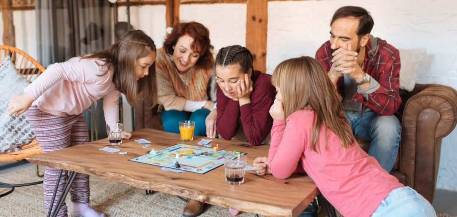Eine Familie spielt in ihrem Wohnzimmer ein Spiel. Neben den beiden Eltern spielen auch drei Mädchen mit. Sie alle sind fröhlich.