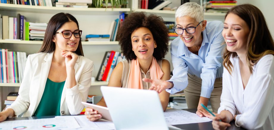 Eine Gruppe von erfolgreichen Business-Frauen arbeitet gemeinsam in einem Büro. Im Hintergrund sieht man ein Bücherregal und im Vordergrund steht ein Laptop.
