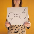 Frau steht vor gelbem Hintergrund. Sie hält ein Schild in die Kamera. Dort sind die beiden Geschlechtszeichen aufgemalt und durch zwei kurze parallele Striche getrennt.
